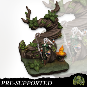 A 3D model of a fantasy character, likely a fairy or elf, standing in a forest setting with a tree and various plants. The character is holding a staff and appears to be in a contemplative or thoughtful pose.