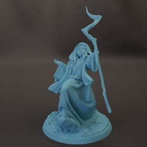 A light blue figurine of a woman holding a staff and standing on a rock.